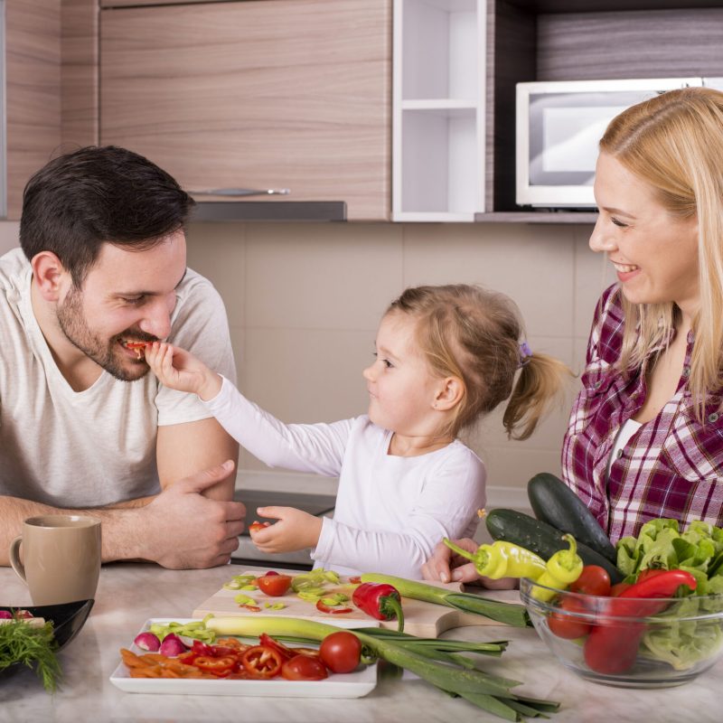 A happy family with their little daughter making a fresh salad with vegetables in the kitchen